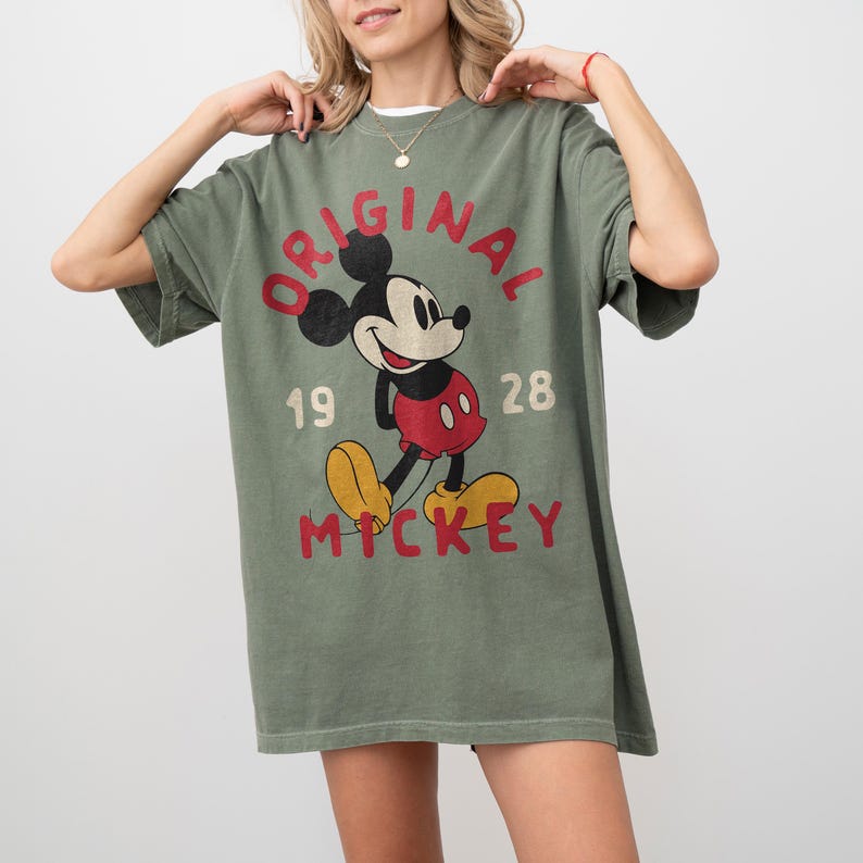 May include: Olive green t-shirt featuring a vintage Mickey Mouse graphic. The design includes the word "ORIGINAL" in red, the numbers "19 28", and the name "MICKEY" in yellow. The shirt has short sleeves and a relaxed fit.