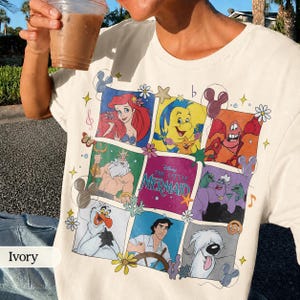 May include: Ivory-colored t-shirt featuring a colorful graphic of characters from Disney's The Little Mermaid. The design includes Ariel, Flounder, Sebastian, King Triton, and Ursula. The shirt has a crew neck and short sleeves.