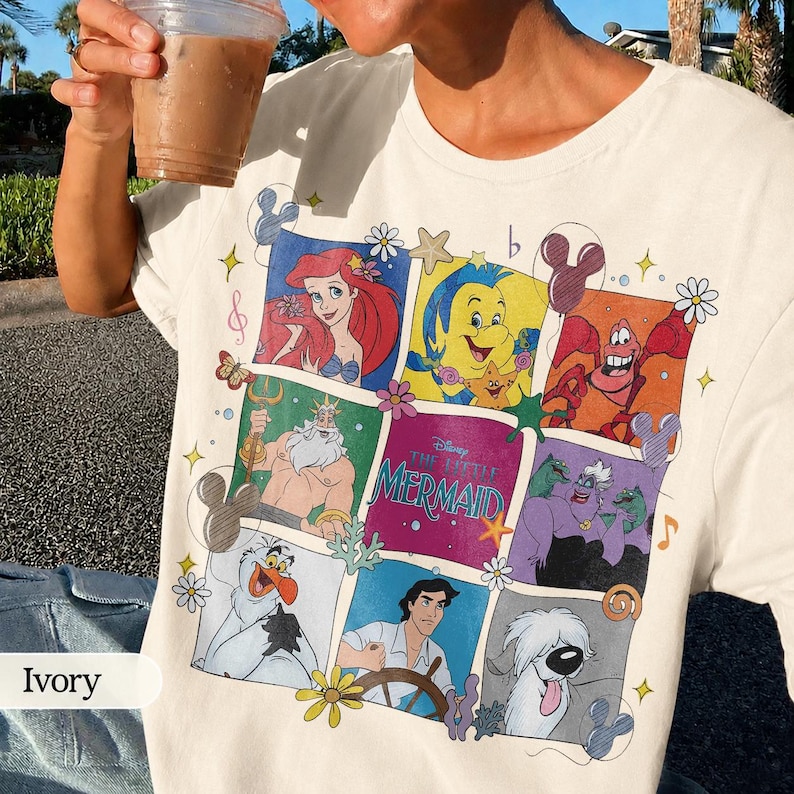May include: Ivory-colored t-shirt featuring a colorful graphic of characters from Disney's The Little Mermaid. The design includes Ariel, Flounder, Sebastian, King Triton, and Ursula. The shirt has a crew neck and short sleeves.