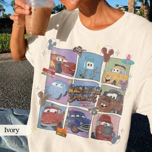 May include: An ivory t-shirt featuring a grid graphic of characters from the movie Cars, including Lightning McQueen, Mater, and Luigi. The design includes the text Radiator Springs and Lightning McQueen, accented with small star and Mickey ear icons.