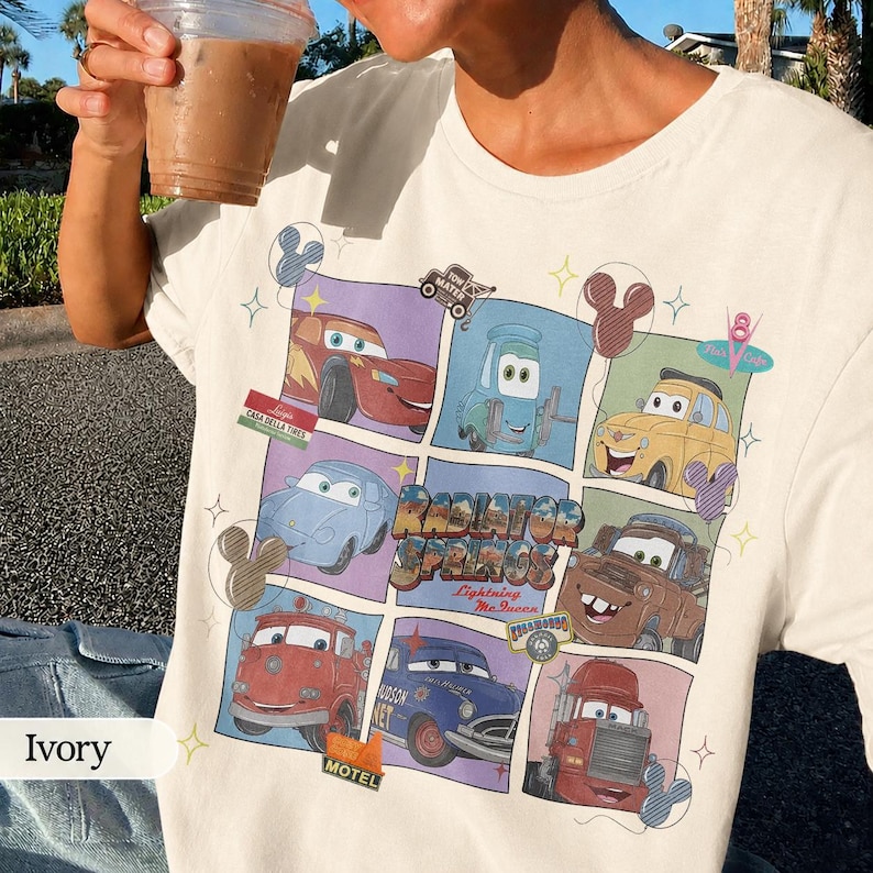 May include: An ivory t-shirt featuring a grid graphic of characters from the movie Cars, including Lightning McQueen, Mater, and Luigi. The design includes the text Radiator Springs and Lightning McQueen, accented with small star and Mickey ear icons.