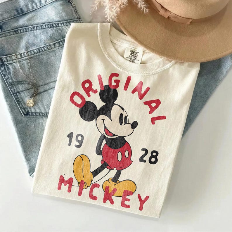 May include: Cream-colored t-shirt featuring a vintage Mickey Mouse graphic. The design includes the word "ORIGINAL" in red, the numbers "1928", and the name "MICKEY" in red. The shirt is folded and placed on top of blue jeans.