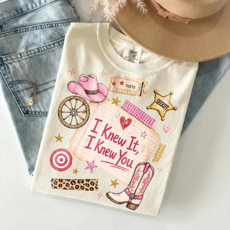 May include: A cream-colored t-shirt featuring a graphic print with a pink cowboy hat, a wagon wheel, a sheriff badge, a cowboy boot, and a notepad with the text "I Knew It, I Knew You" surrounded by stars, hearts, and decorative tape patterns.