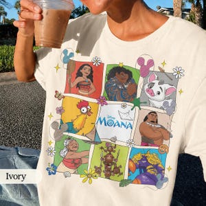 Op de afbeelding: Ivoor kleurig T-shirt met een kleurrijke afbeelding van personages uit de Disney film "Moana". Het ontwerp bevat afbeeldingen van Moana, Maui, Hei Hei, Pua en anderen, met het logo van de film in het midden. Het shirt is gemaakt van zacht materiaal.