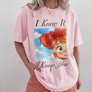 K&ouml;nnte beinhalten: Ein hellrosa T-Shirt mit kurzen &Auml;rmeln und einem Grafikdruck einer Zeichentrickfigur mit roten Haaren und rotem Hut vor einem blauen Himmel mit Wolken. Der Text lautet I Knew It EST 2026 &uuml;ber dem Bild und I Knew You mit einem Violinschl&uuml;ssel.