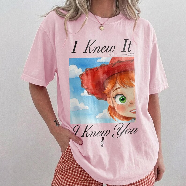 K&ouml;nnte beinhalten: Ein hellrosa T-Shirt mit kurzen &Auml;rmeln und einem Grafikdruck einer Zeichentrickfigur mit roten Haaren und rotem Hut vor einem blauen Himmel mit Wolken. Der Text lautet I Knew It EST 2026 &uuml;ber dem Bild und I Knew You mit einem Violinschl&uuml;ssel.