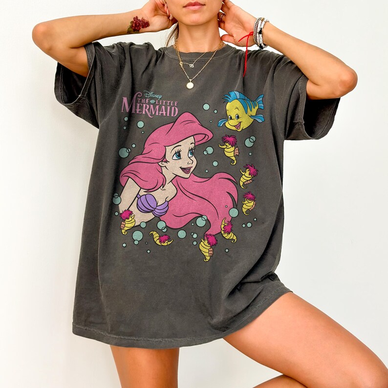 May include: Dark gray t-shirt featuring a graphic of Ariel from Disney's The Little Mermaid. The design includes Ariel with pink hair, a purple seashell bra, and the text "Disney The Little Mermaid". Also includes Flounder and seahorse characters.
