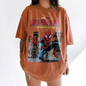 May include: An oversized burnt orange t-shirt featuring a graphic print of Spider-Man in a dynamic pose against a city skyline. The text reads Marvel Studios Spider-Man Brand New Day. The shirt has short sleeves and a crew neckline.