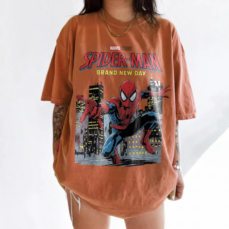 May include: An oversized burnt orange t-shirt featuring a graphic print of Spider-Man in a dynamic pose against a city skyline. The text reads Marvel Studios Spider-Man Brand New Day. The shirt has short sleeves and a crew neckline.