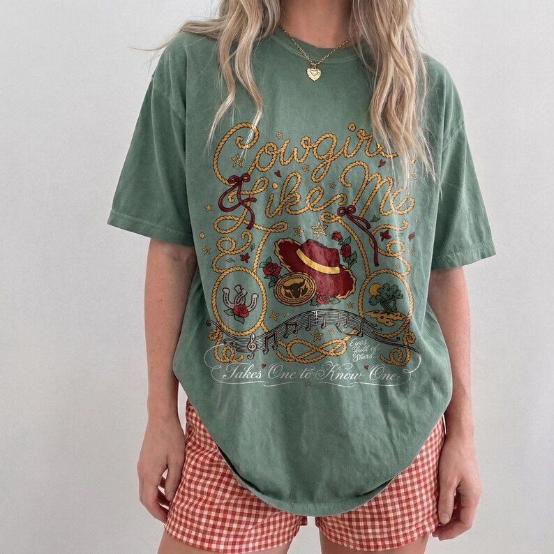 May include: A sage green short-sleeve t-shirt featuring a graphic print with yellow rope lettering that reads Cowgirls Like Me and Takes One to Know One. The design includes a red cowboy hat, a gold belt buckle, roses, and musical notes.