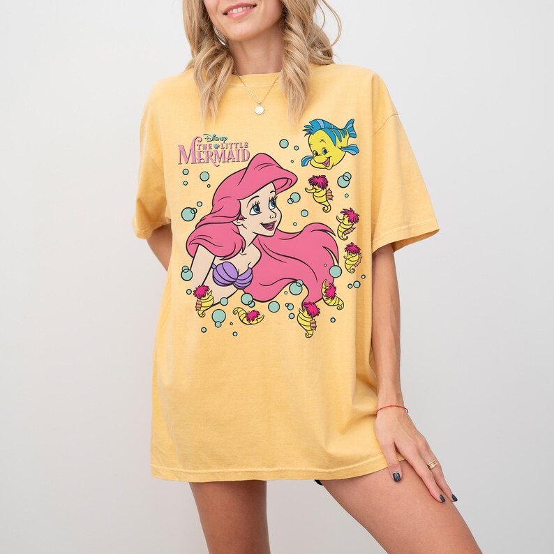 May include: Yellow t-shirt featuring a graphic of Ariel from Disney's The Little Mermaid. Ariel has pink hair and a purple seashell top. The shirt also includes the character Flounder and the words "The Little Mermaid".