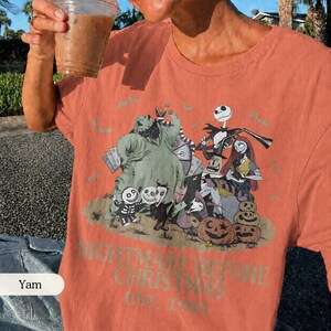 May include: Coral-colored t-shirt featuring a graphic from "The Nightmare Before Christmas." The design includes characters like Jack Skellington, Sally, and Oogie Boogie, along with pumpkins. The text "NIGHTMARE BEFORE CHRISTMAS EST. 1993" is printed below the graphic.