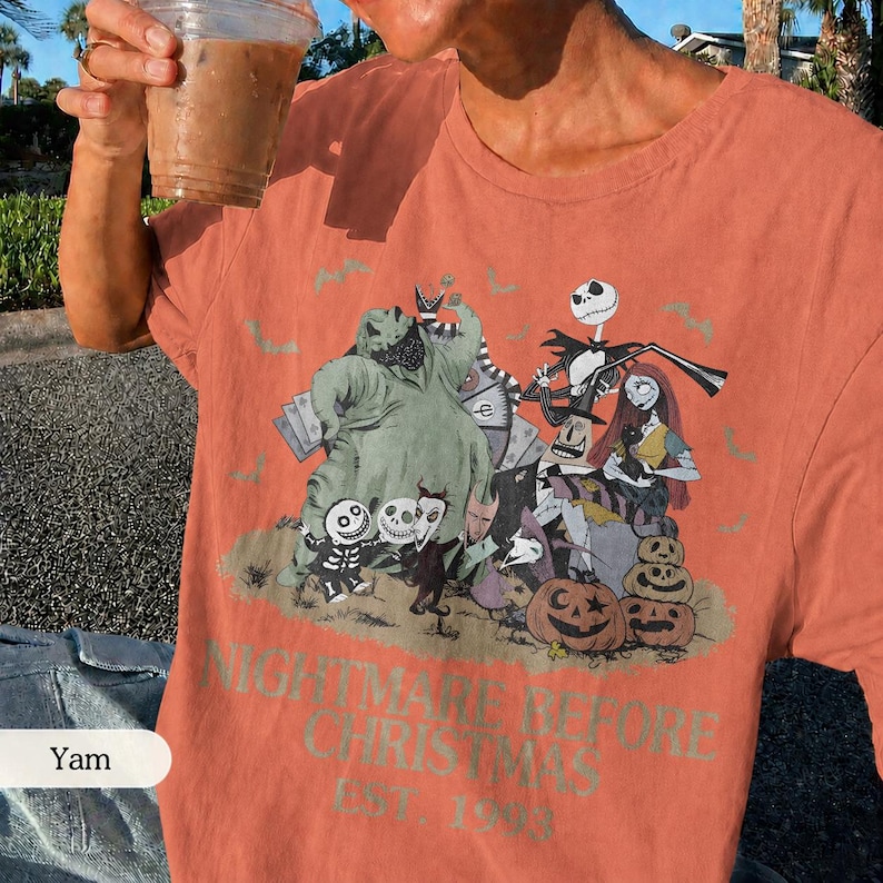 May include: Coral-colored t-shirt featuring a graphic from "The Nightmare Before Christmas." The design includes characters like Jack Skellington, Sally, and Oogie Boogie, along with pumpkins. The text "NIGHTMARE BEFORE CHRISTMAS EST. 1993" is printed below the graphic.