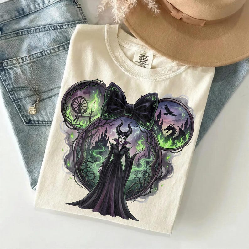May include: Cream-colored t-shirt featuring a graphic of a Disney villain in a black dress, surrounded by a green and purple forest scene. The design is shaped like Mickey Mouse ears with a black bow. The shirt is folded on a light blue surface.