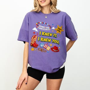 May include: A purple short-sleeve t-shirt featuring a graphic print with the text "I KNEW IT, I KNEW YOU," a beaded bracelet, a yellow sheriff star badge with "INFINITY AND BEYOND," red cowboy boots, blue and white blocks with "TS5," and a hat.