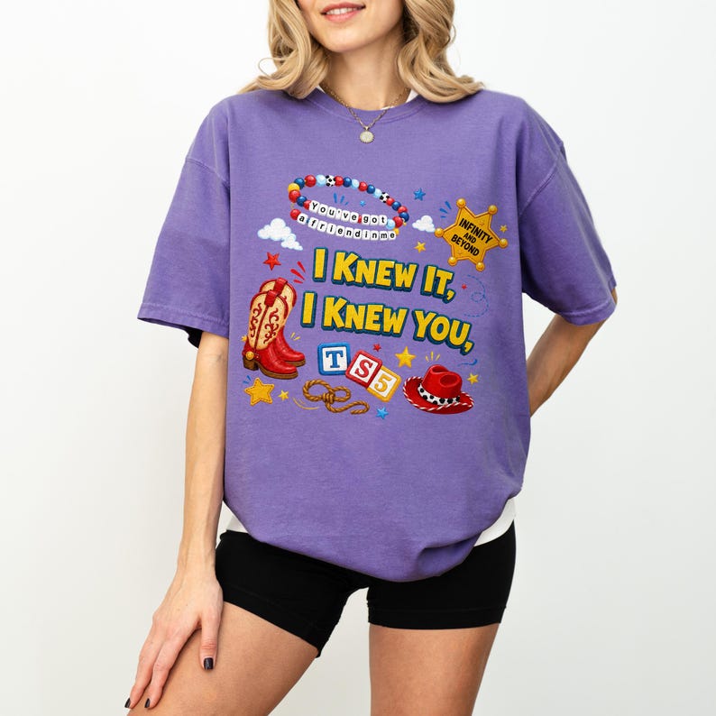 May include: A purple short-sleeve t-shirt featuring a graphic print with the text "I KNEW IT, I KNEW YOU," a beaded bracelet, a yellow sheriff star badge with "INFINITY AND BEYOND," red cowboy boots, blue and white blocks with "TS5," and a hat.
