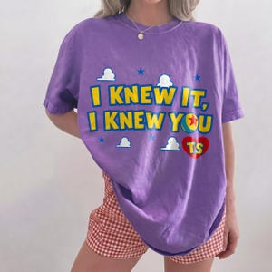 May include: A purple short-sleeve t-shirt featuring the yellow text I KNEW IT, I KNEW YOU with a red heart containing the letters TS. The design includes small white clouds and blue stars, with the letter O replaced by a colorful ball graphic.