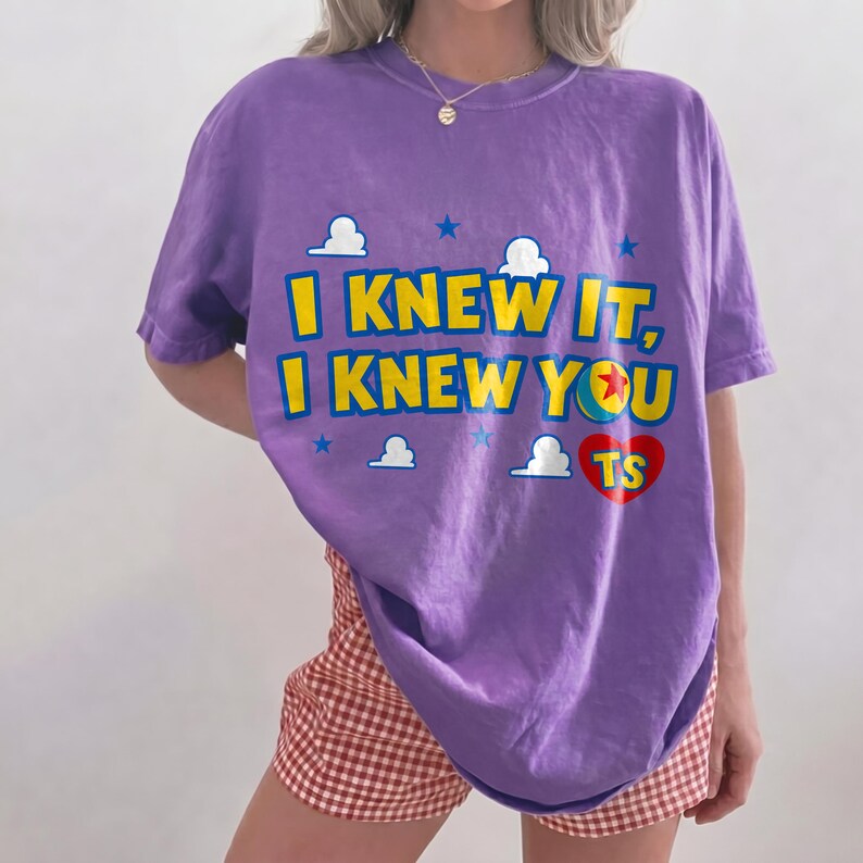 May include: A purple short-sleeve t-shirt featuring the yellow text I KNEW IT, I KNEW YOU with a red heart containing the letters TS. The design includes small white clouds and blue stars, with the letter O replaced by a colorful ball graphic.