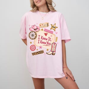 May include: A light pink short-sleeve t-shirt featuring a graphic print of a pink cowboy hat, a wagon wheel, a sheriff badge, a cowboy boot, and a notepad with the text "I Knew It, I Knew You" written in red, surrounded by small stars and hearts.