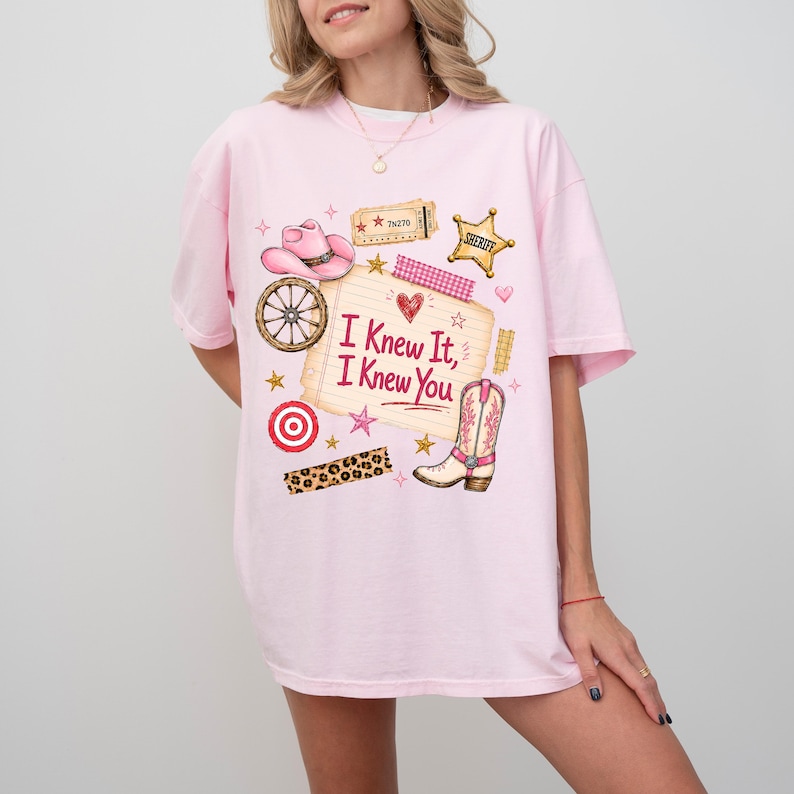 May include: A light pink short-sleeve t-shirt featuring a graphic print of a pink cowboy hat, a wagon wheel, a sheriff badge, a cowboy boot, and a notepad with the text "I Knew It, I Knew You" written in red, surrounded by small stars and hearts.