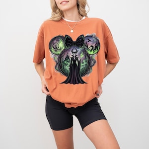 May include: An orange t-shirt featuring a graphic of a Disney villain in a black dress, with a black bow and mouse ears. The design includes a castle, a spinning wheel, and a dragon, with green and black accents.