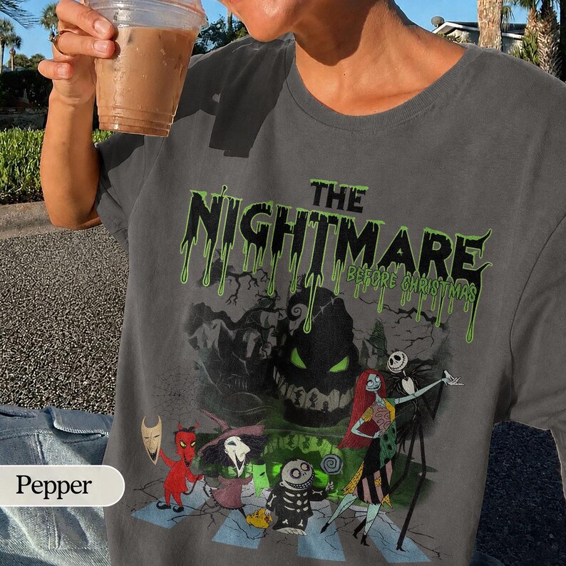 K&ouml;nnte beinhalten: Graues T-Shirt mit dem Text "THE NIGHTMARE BEFORE CHRISTMAS" in tropfenden gr&uuml;nen Buchstaben. Die Grafik zeigt Charaktere aus dem Film. Das T-Shirt hat einen Rundhalsausschnitt.