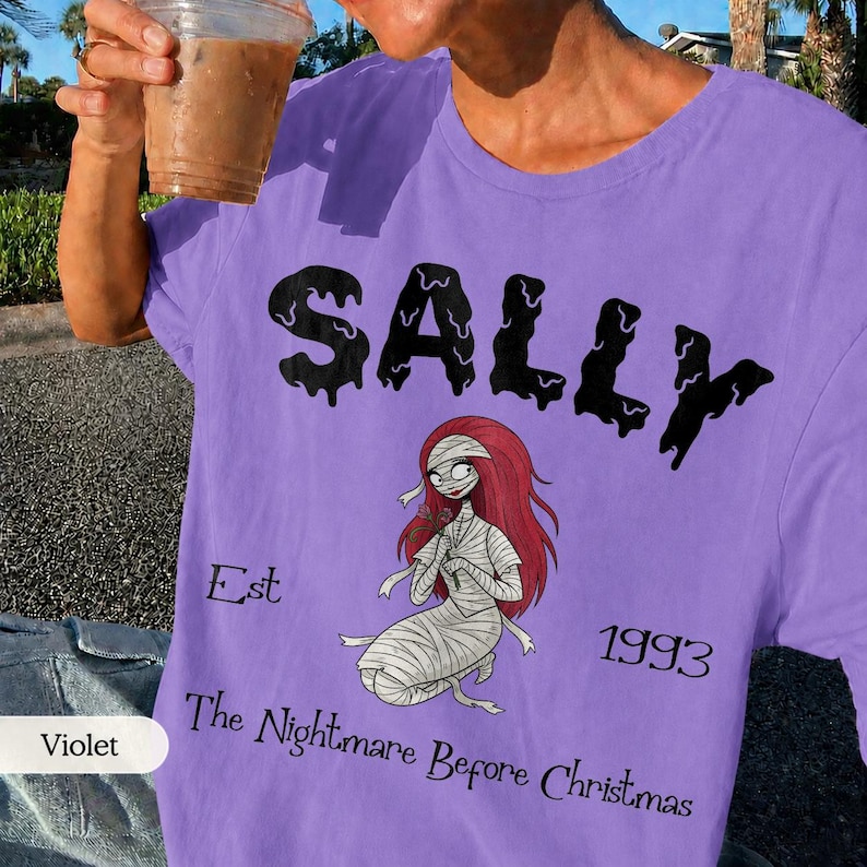 May include: Violet-colored t-shirt featuring the name "SALLY" in black dripping font, with an illustration of Sally from "The Nightmare Before Christmas." The shirt also includes the text "Est 1993" and "The Nightmare Before Christmas."