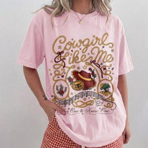 May include: A light pink short-sleeve t-shirt featuring a graphic print with the text Cowgirl Like Me, Eyes full of Stars, and Takes One to Know One. The design includes a brown cowboy hat, a horseshoe, a rope border, and musical notes.