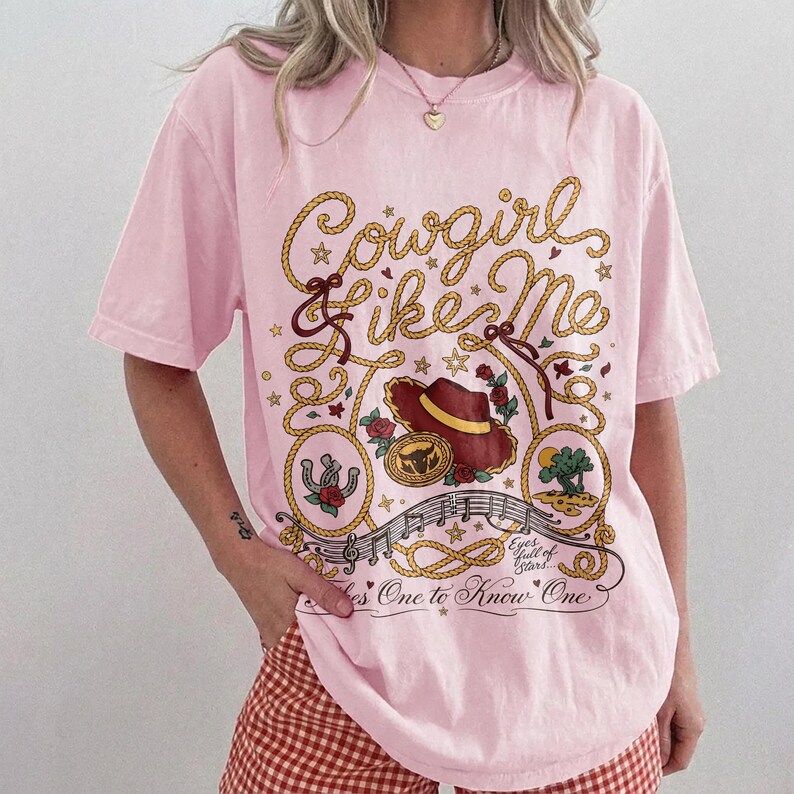 May include: A light pink short-sleeve t-shirt featuring a graphic print with the text Cowgirl Like Me, Eyes full of Stars, and Takes One to Know One. The design includes a brown cowboy hat, a horseshoe, a rope border, and musical notes.