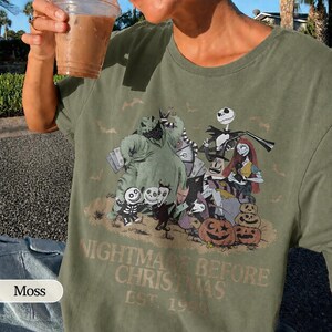 May include: Moss green t-shirt featuring a graphic from "The Nightmare Before Christmas." The design includes characters like Jack Skellington, Sally, and Oogie Boogie, along with pumpkins. The text "NIGHTMARE BEFORE CHRISTMAS EST. 1993" is printed below the characters.