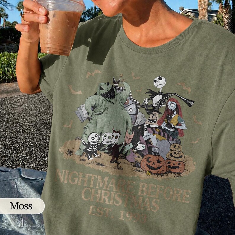 May include: Moss green t-shirt featuring a graphic from "The Nightmare Before Christmas." The design includes characters like Jack Skellington, Sally, and Oogie Boogie, along with pumpkins. The text "NIGHTMARE BEFORE CHRISTMAS EST. 1993" is printed below the characters.