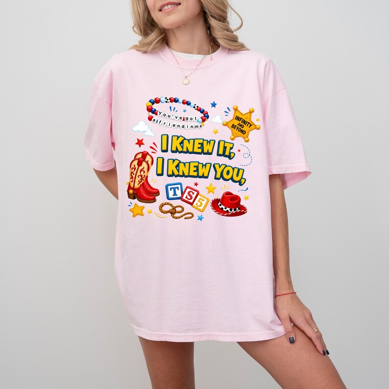May include: A light pink t-shirt featuring a graphic print with the text I KNEW IT, I KNEW YOU, TS5, and INFINITY AND BEYOND. The design includes a beaded bracelet, a yellow sheriff star, red cowboy boots, blue and red blocks, and a red hat.