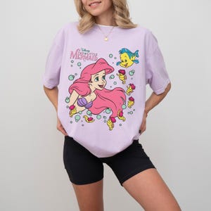 May include: A light purple t-shirt featuring a graphic of Ariel from Disney's The Little Mermaid. The design includes Ariel with pink hair, a purple seashell top, and Flounder. The shirt has the text "The Little Mermaid".