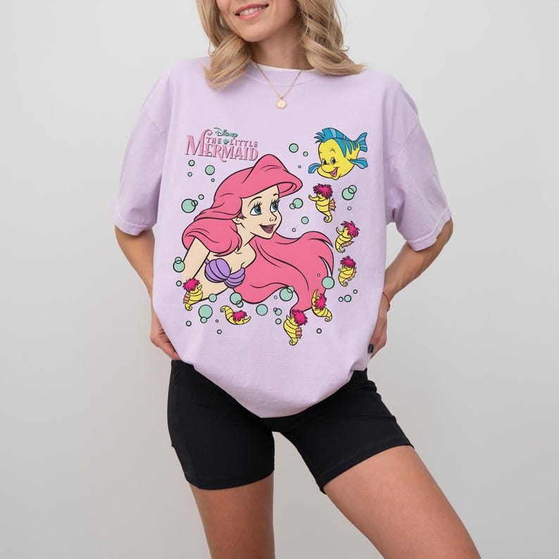 May include: A light purple t-shirt featuring a graphic of Ariel from Disney's The Little Mermaid. The design includes Ariel with pink hair, a purple seashell top, and Flounder. The shirt has the text "The Little Mermaid".
