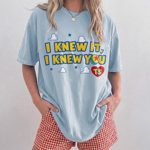 May include: A light blue short-sleeve t-shirt featuring the yellow text I KNEW IT, I KNEW YOU with a red heart containing the letters TS and a small ball graphic. The design includes small blue stars and white cloud shapes printed on the front.