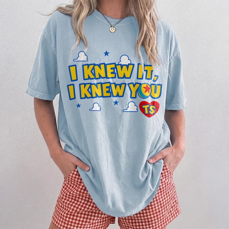 May include: A light blue short-sleeve t-shirt featuring the yellow text I KNEW IT, I KNEW YOU with a red heart containing the letters TS and a small ball graphic. The design includes small blue stars and white cloud shapes printed on the front.