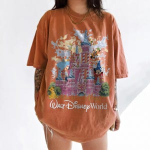 May include: An oversized burnt orange t-shirt featuring a graphic print of the Walt Disney World 25th anniversary castle. The design includes various animated characters, a large number 25, and the white text Walt Disney World at the bottom.