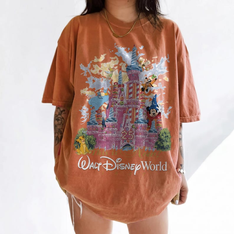 May include: An oversized burnt orange t-shirt featuring a graphic print of the Walt Disney World 25th anniversary castle. The design includes various animated characters, a large number 25, and the white text Walt Disney World at the bottom.