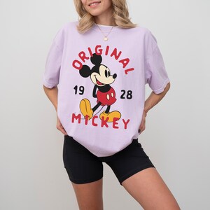 May include: A light purple t-shirt featuring a classic Mickey Mouse graphic. The design includes the words "ORIGINAL MICKEY" in red, along with the numbers "19" and "28". Mickey Mouse is depicted in his iconic red shorts, yellow shoes, and black gloves.