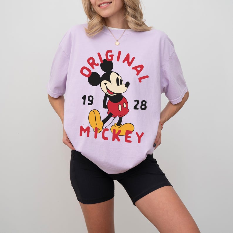 May include: A light purple t-shirt featuring a classic Mickey Mouse graphic. The design includes the words "ORIGINAL MICKEY" in red, along with the numbers "19" and "28". Mickey Mouse is depicted in his iconic red shorts, yellow shoes, and black gloves.