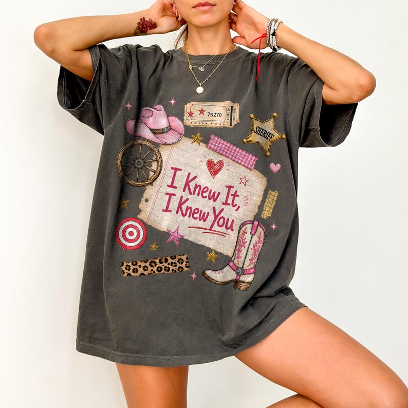 May include: A charcoal gray t-shirt featuring a graphic print of a pink cowboy hat, a sheriff badge, a wagon wheel, a leopard print strip, and a pink cowboy boot. The center text reads "I Knew It, I Knew You" on a paper-like background.