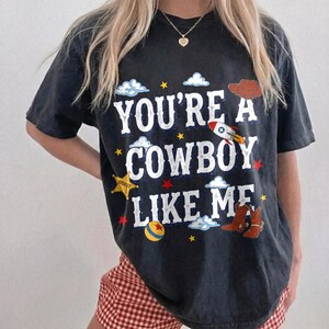 Puede incluir: Una camiseta gris carb&oacute;n de manga corta con el texto blanco "YOU'RE A COWBOY LIKE ME" rodeado de gr&aacute;ficos coloridos que incluyen un sombrero de vaquero marr&oacute;n, un cohete, una estrella de sheriff amarilla, una pelota roja y amarilla y botas marrones.