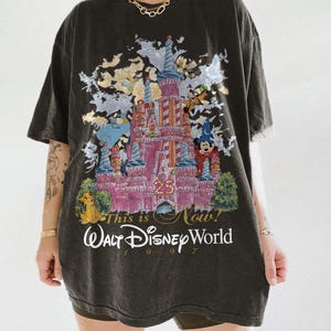 May include: A black short-sleeve t-shirt featuring a graphic of the Walt Disney World 25th anniversary castle. The print includes characters like Mickey Mouse, Simba, and Tinker Bell, with the text This is Now! Walt Disney World 1997.