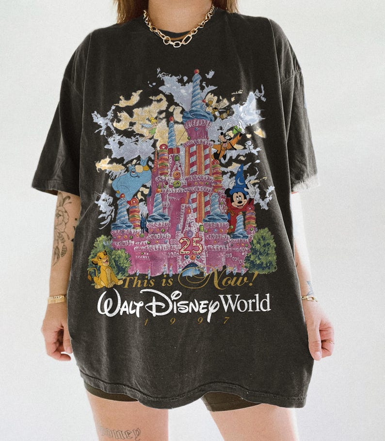 May include: A black short-sleeve t-shirt featuring a graphic of the Walt Disney World 25th anniversary castle. The print includes characters like Mickey Mouse, Simba, and Tinker Bell, with the text This is Now! Walt Disney World 1997.
