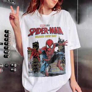 May include: A white short-sleeve t-shirt featuring a graphic print of Spider-Man in a dynamic pose against a city skyline. The text reads Marvel Studios Spider-Man Brand New Day. The design includes red, blue, and yellow colors on the fabric.