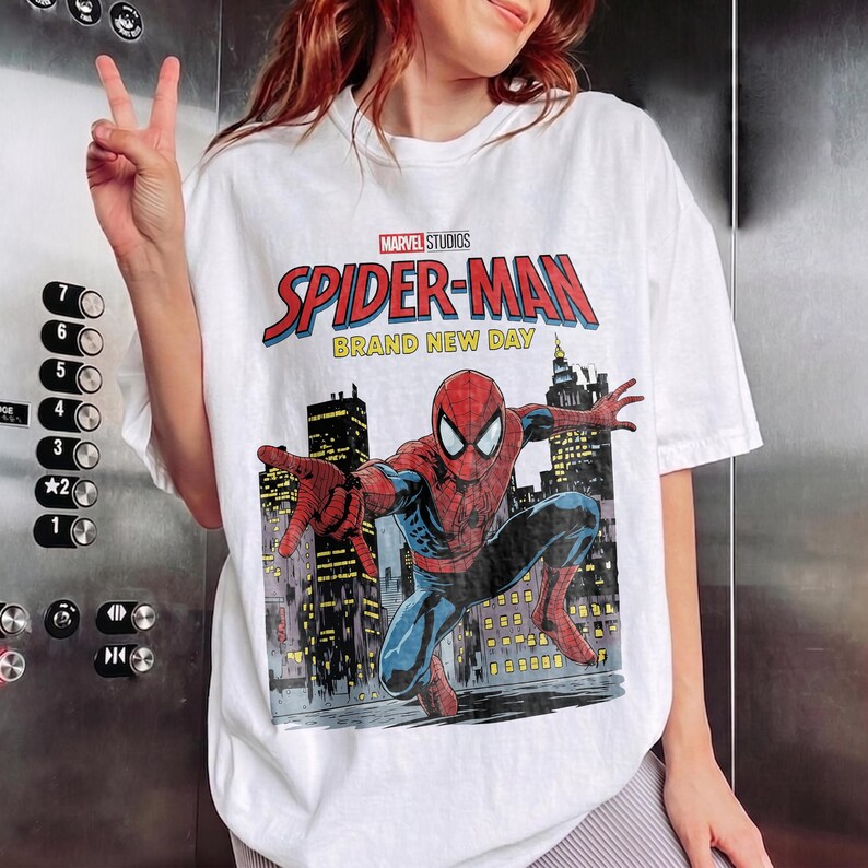 May include: A white short-sleeve t-shirt featuring a graphic print of Spider-Man in a dynamic pose against a city skyline. The text reads Marvel Studios Spider-Man Brand New Day. The design includes red, blue, and yellow colors on the fabric.