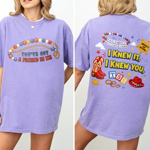 Pu&ograve; includere: Una t-shirt viola chiaro con una grafica sul davanti e sul retro. Il davanti mostra una collana di perline e il testo "You've got a friend in me". Il retro mostra perline, una stella da sceriffo, stivali da cowboy, blocchi e un cappello.
