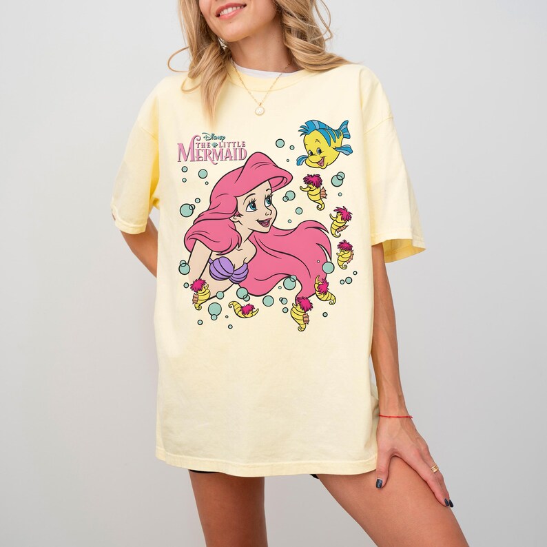 May include: A light yellow t-shirt featuring a graphic of Ariel from Disney's The Little Mermaid. Ariel has long pink hair and a purple seashell bra. The shirt also features Flounder and other sea creatures. The text "Disney The Little Mermaid" is above the graphic.