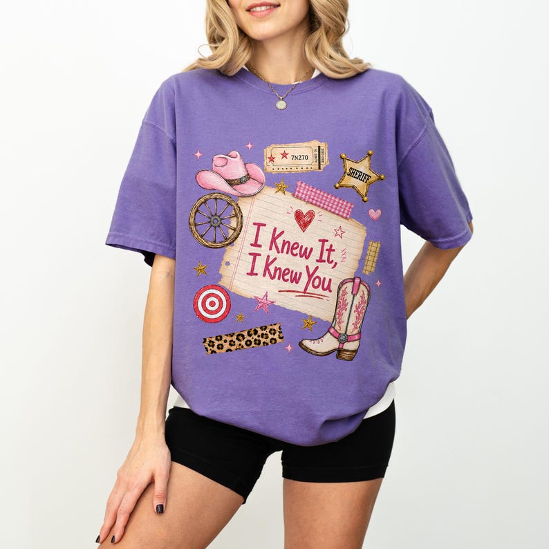 May include: A purple short-sleeve t-shirt featuring a graphic print of a pink cowboy hat, a wagon wheel, a sheriff star, a leopard print strip, and a cowboy boot. The center shows a notepad with the text "I Knew It, I Knew You" in red.