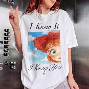 K&ouml;nnte beinhalten: Ein wei&szlig;es T-Shirt mit kurzen &Auml;rmeln und einem Grafikdruck einer Zeichentrickfigur mit roten Haaren und einem roten Hut vor einem blauen Himmel mit Wolken. Der Text lautet I Knew It EST 2026 oben und I Knew You mit einem Violinschl&uuml;ssel unten.